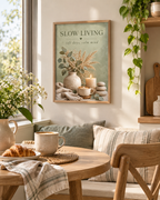 Slow Living artwork with sage green foliage, dried grasses, a coffee cup, candle, ceramic vase and pebbles styled in a window seat