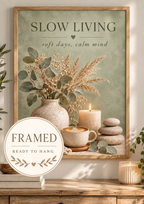 Slow Living artwork with sage green foliage, dried grasses, a coffee cup, candle, ceramic vase and pebbles; framed and ready to hang badge