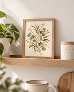 Delicate sage green eucalyptus, olive-toned foliage and ivory-white flowers on a warm neutral background styled in a shelf display